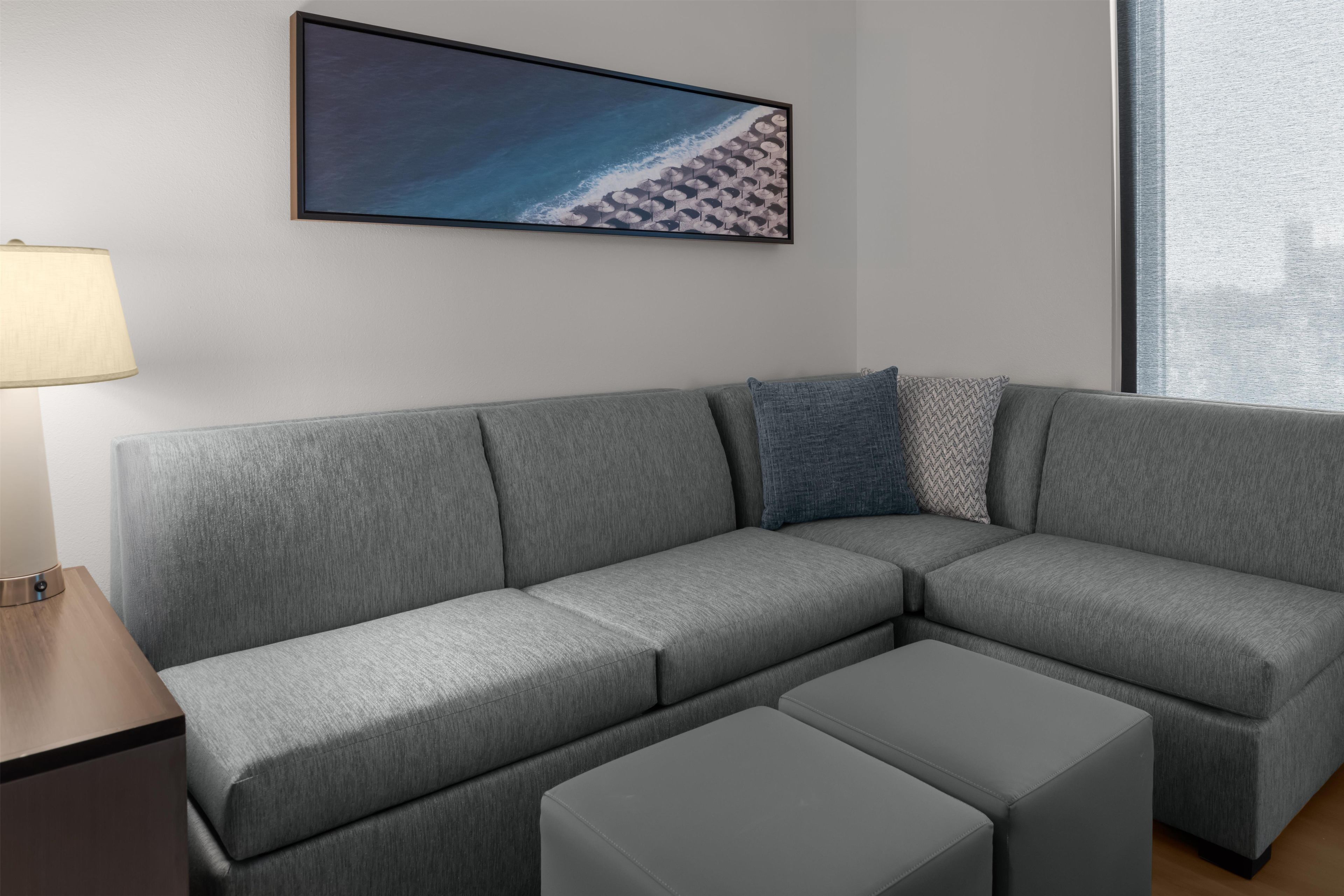 A grey sectional sofa with a matching ottoman, a framed picture of a beach, and a lamp on a wooden side table.