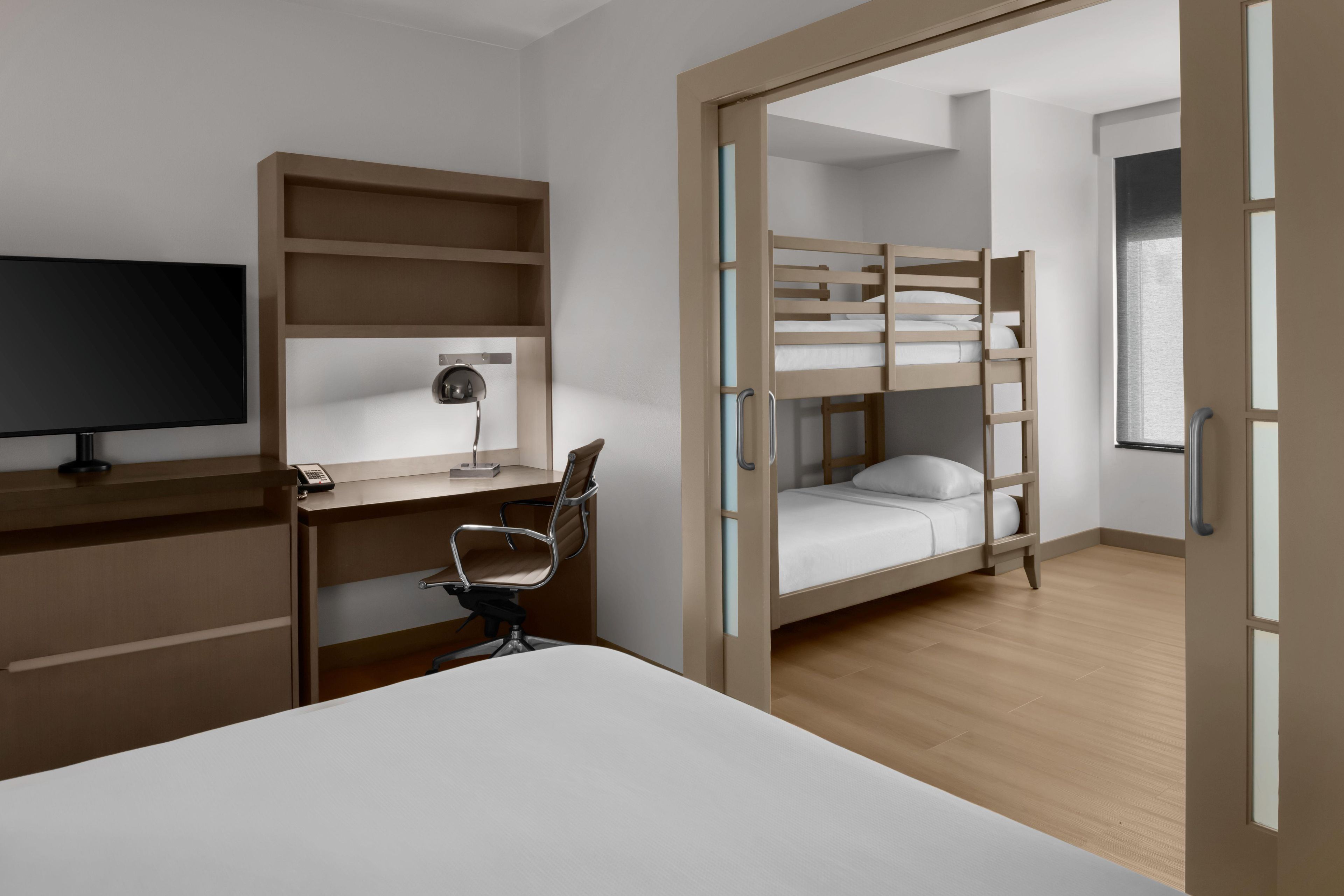 A room with a bunk bed, a desk with a chair, a television, and a desk lamp.