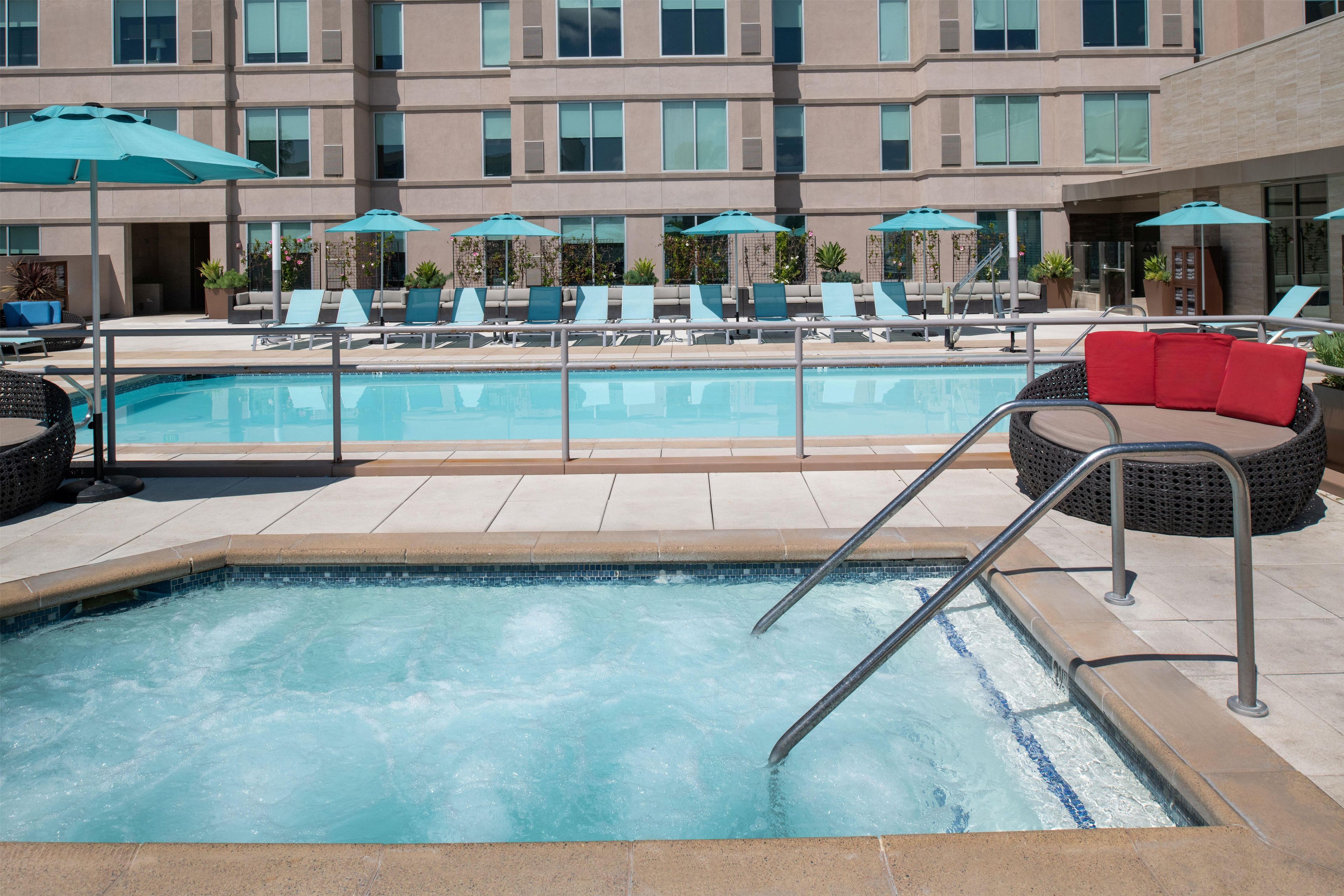 A swimming pool with lounge chairs and umbrellas, surrounded by a building with multiple floors and windows.