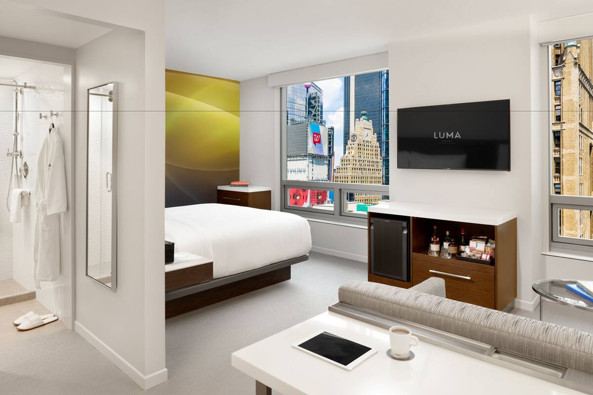 A modern hotel room with a bed, a sofa, a TV, and a window with a city view.
