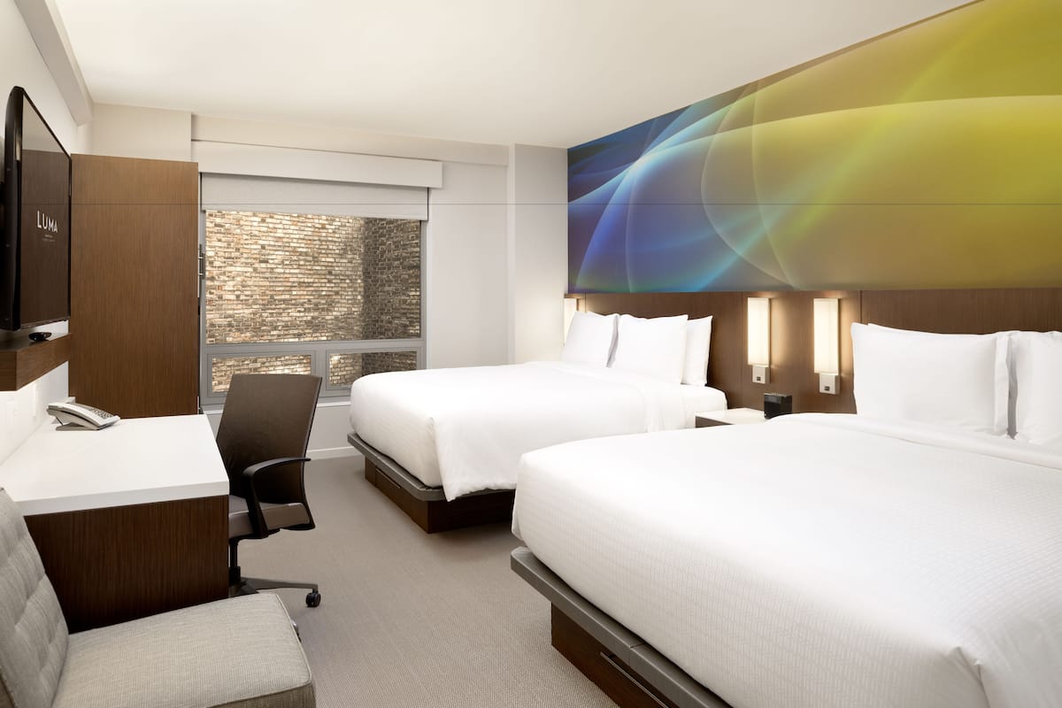 A hotel room with two beds, a desk, a chair, a TV, and a large window with a brick wall view.