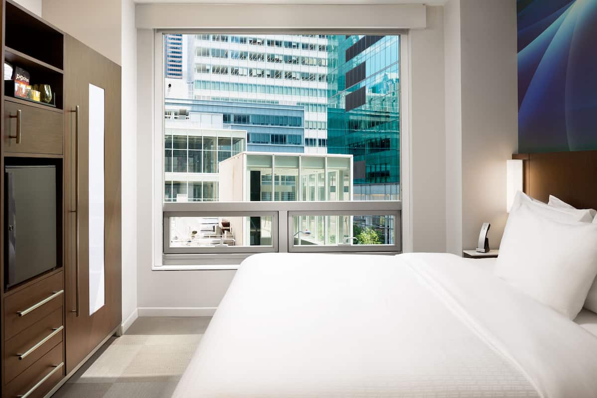 A modern hotel room with a large bed, a wardrobe, and a window offering a view of a cityscape.