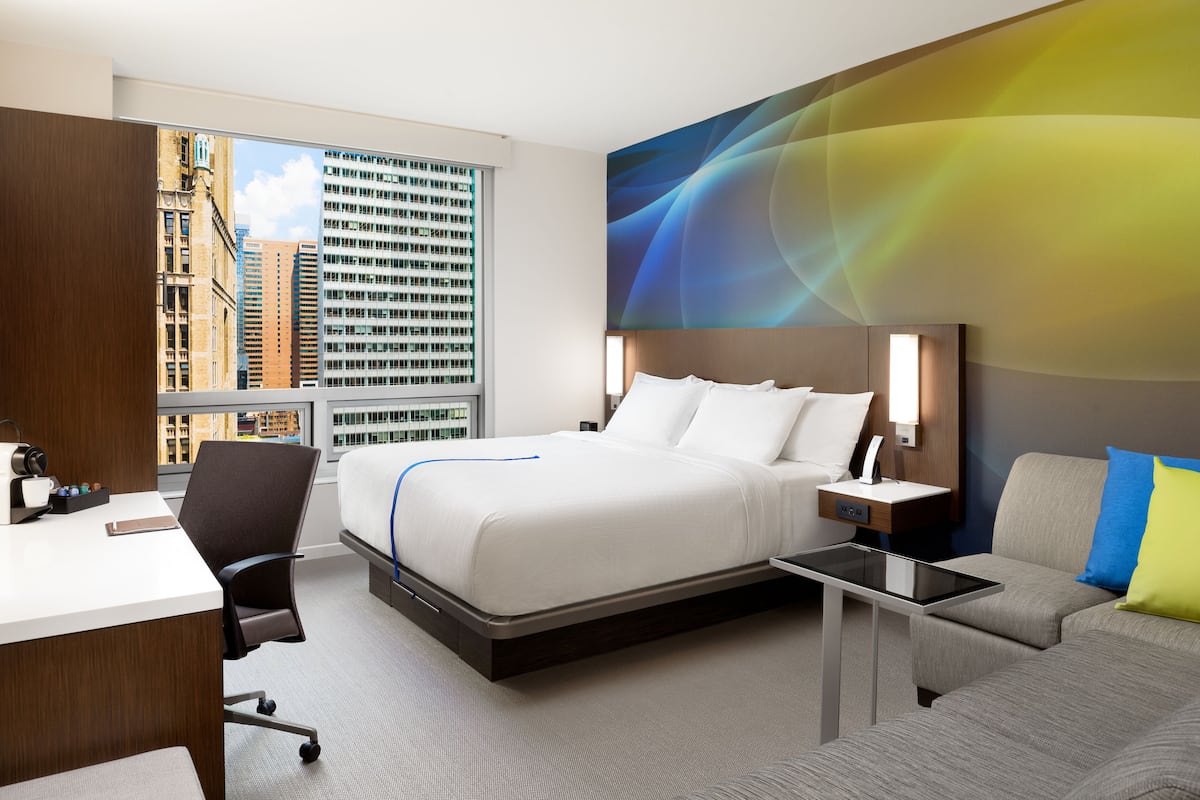A hotel room with a bed, desk, chair, and a view of city buildings.