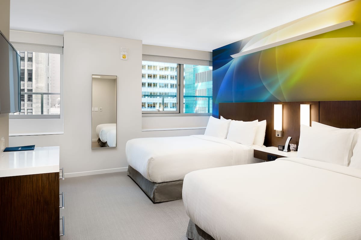 A modern hotel room with two beds, a large window, a mirror, and a wall-mounted light fixture.