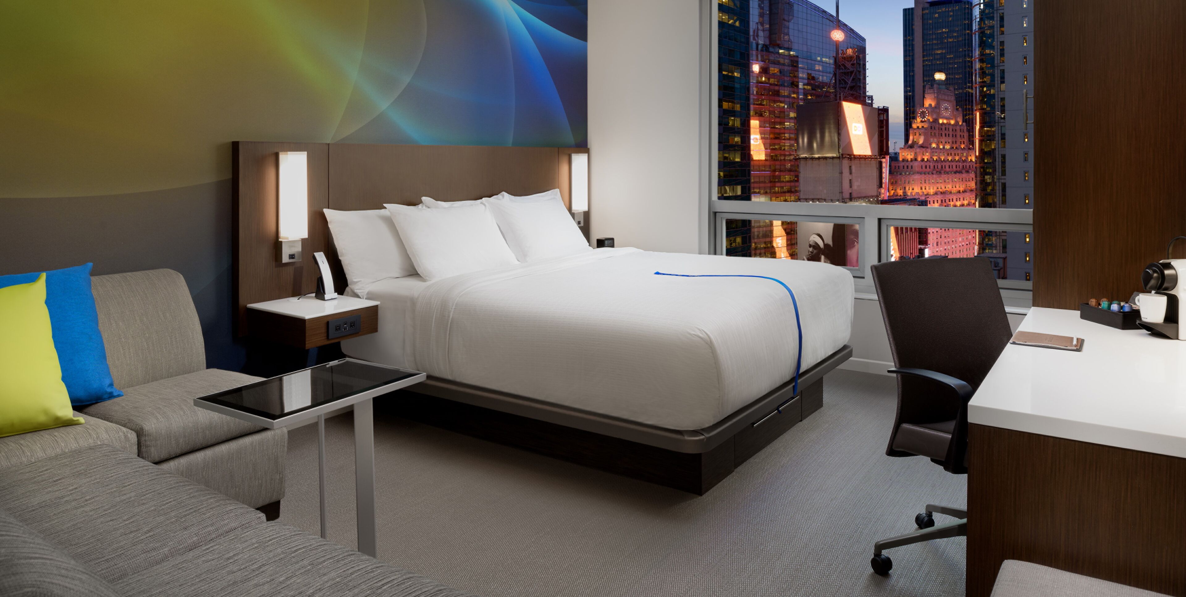 Habitación de hotel con cama, mesita de noche, escritorio y vista a la ciudad.