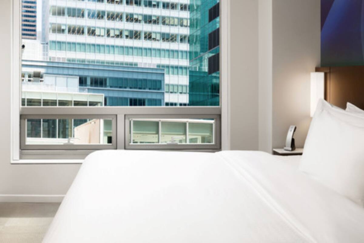 A hotel room with a large bed, a bedside table with a lamp, and a window offering a view of a modern building.