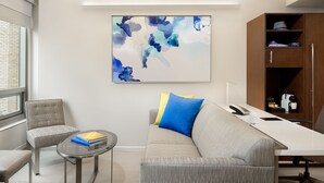 A modern living room with a grey sofa, a small round table, and a chair. There is a painting on the wall and a wooden cabinet with shelves. - LUMA Hotel Times Square (New York)