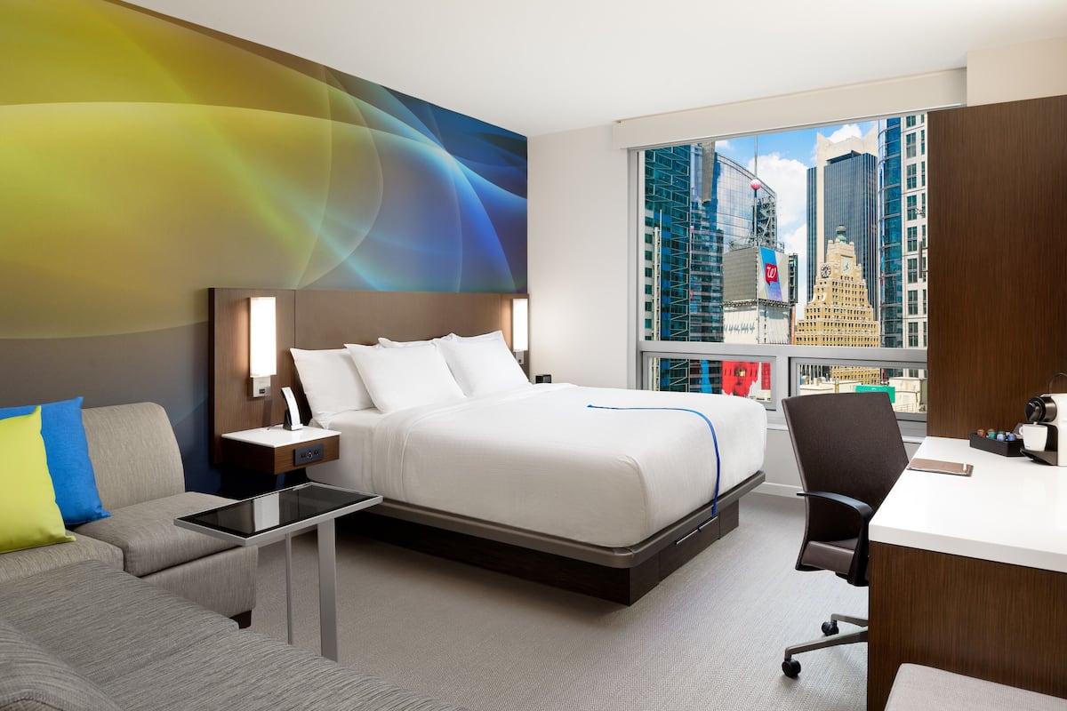 A modern hotel room with a bed, a desk, a chair, a sofa, and a large window with a city view.