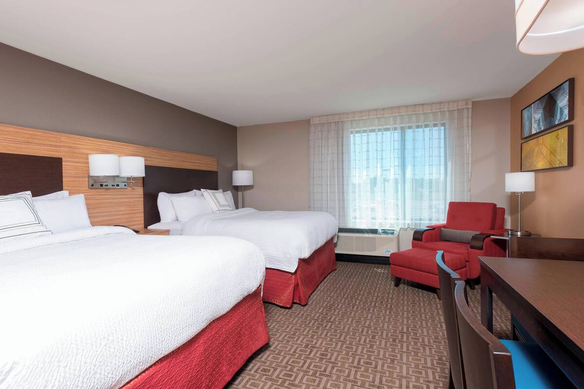 A hotel room with two beds, a red armchair, a wooden desk, and a large window with sheer curtains.