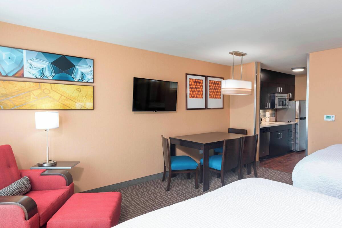 A hotel room with a bed, a desk, a chair, a TV, and a kitchenette.