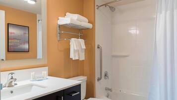 Een moderne badkamer met een wit wastafelmeubel, toilet en een douche met glazen wand. Handdoeken liggen op een plank en er hangt een afbeelding in een lijst aan de muur.