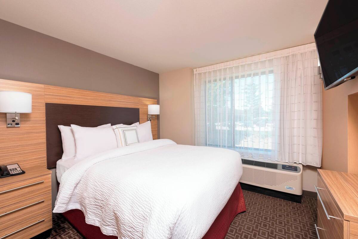 A hotel room with a large bed, two bedside tables, a flat-screen TV, an air conditioning unit, and a window with curtains.