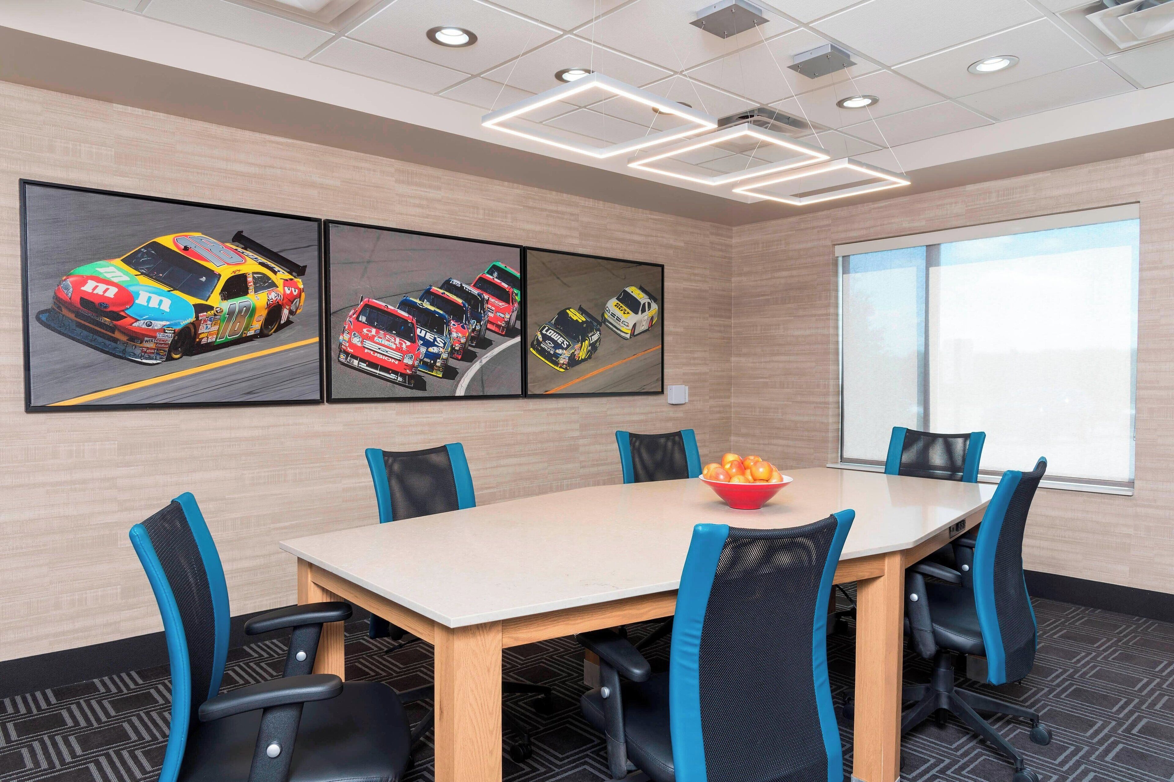 Een vergaderruimte met een lange tafel, zes stoelen en drie grote posters van raceauto's aan de muur.