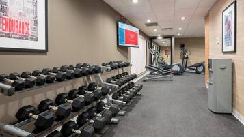 Un gimnasio con filas de mancuernas, cintas de correr y una pantalla digital mostrando contenido de acondicionamiento físico.