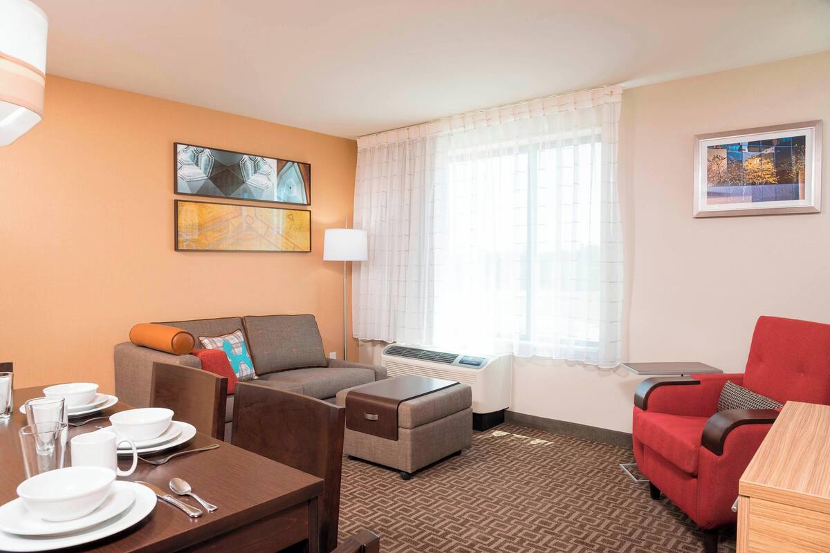 A hotel room with a dining table set for four, a sofa, a red armchair, and a small table with a lamp.