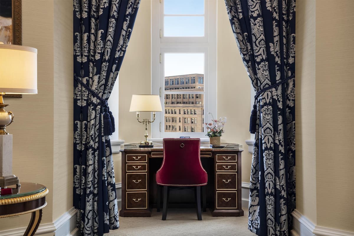 Ein Zimmer mit Schreibtisch, Stuhl und Lampe. Ein Fenster mit Vorhängen bietet Blick auf umliegende Gebäude.