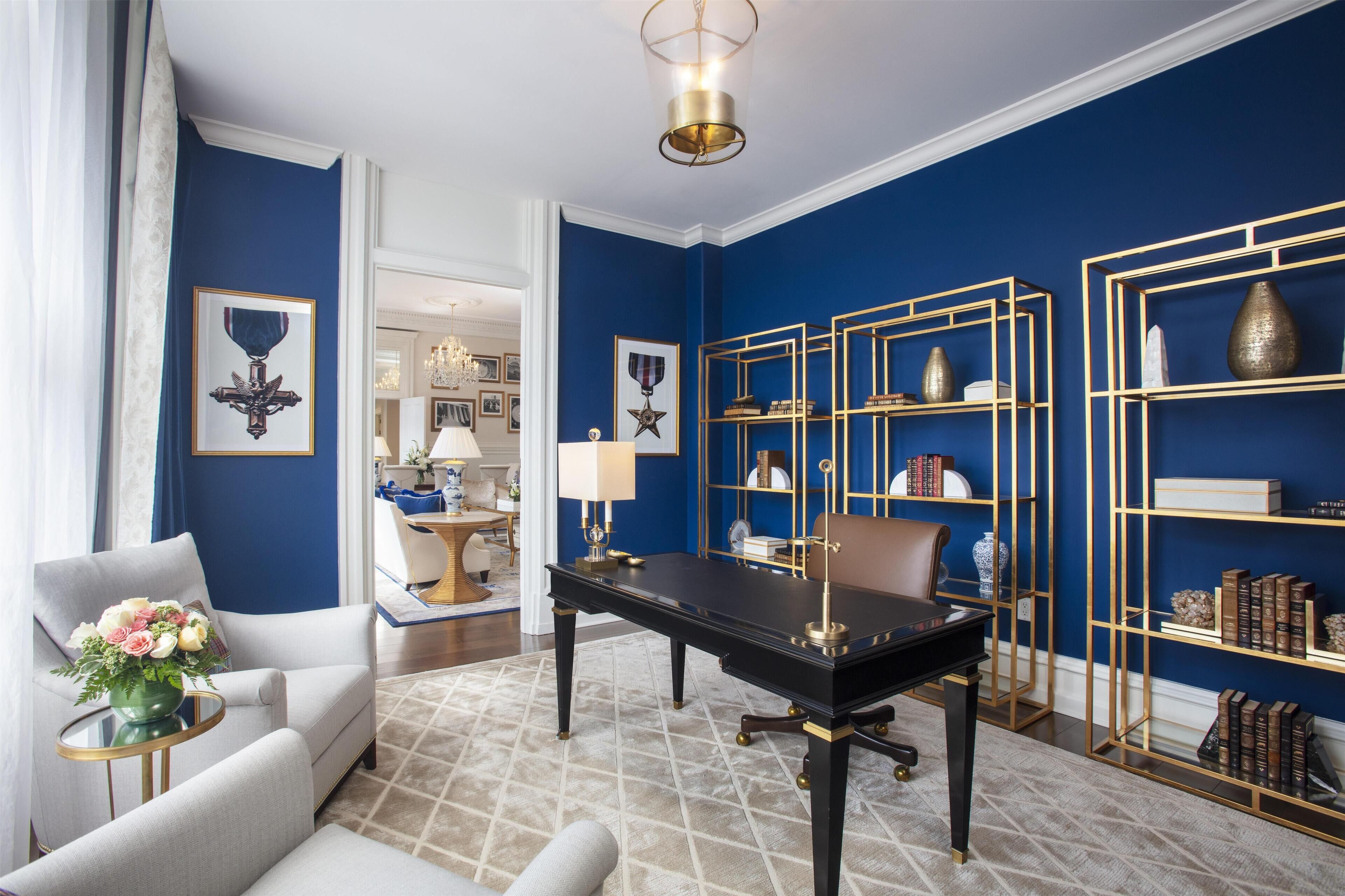 A room with a blue accent wall, a black desk, a white armchair, and a gold-framed bookshelf.