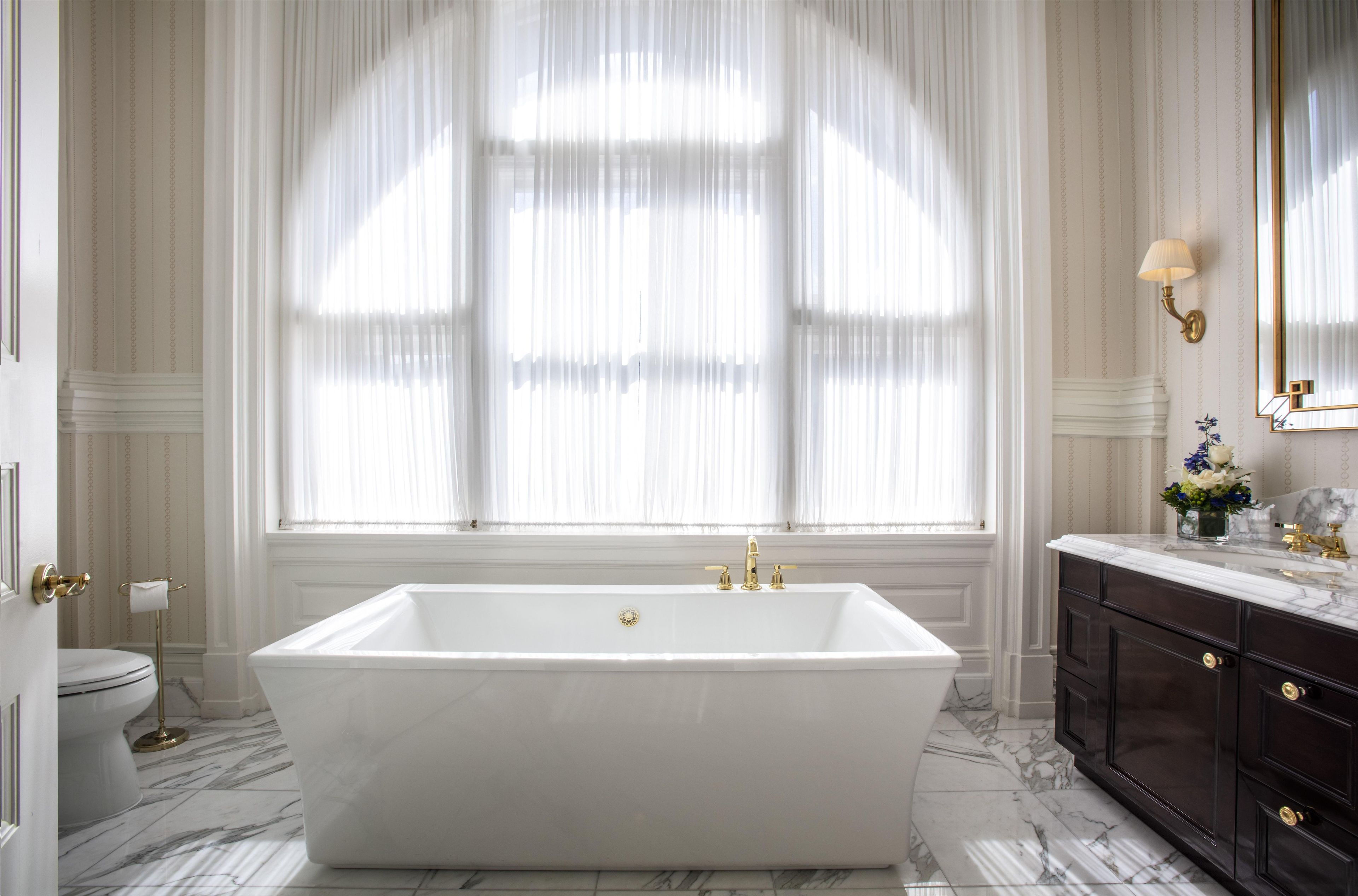 A bathroom with a freestanding bathtub, a marble floor, and a large window with sheer curtains.