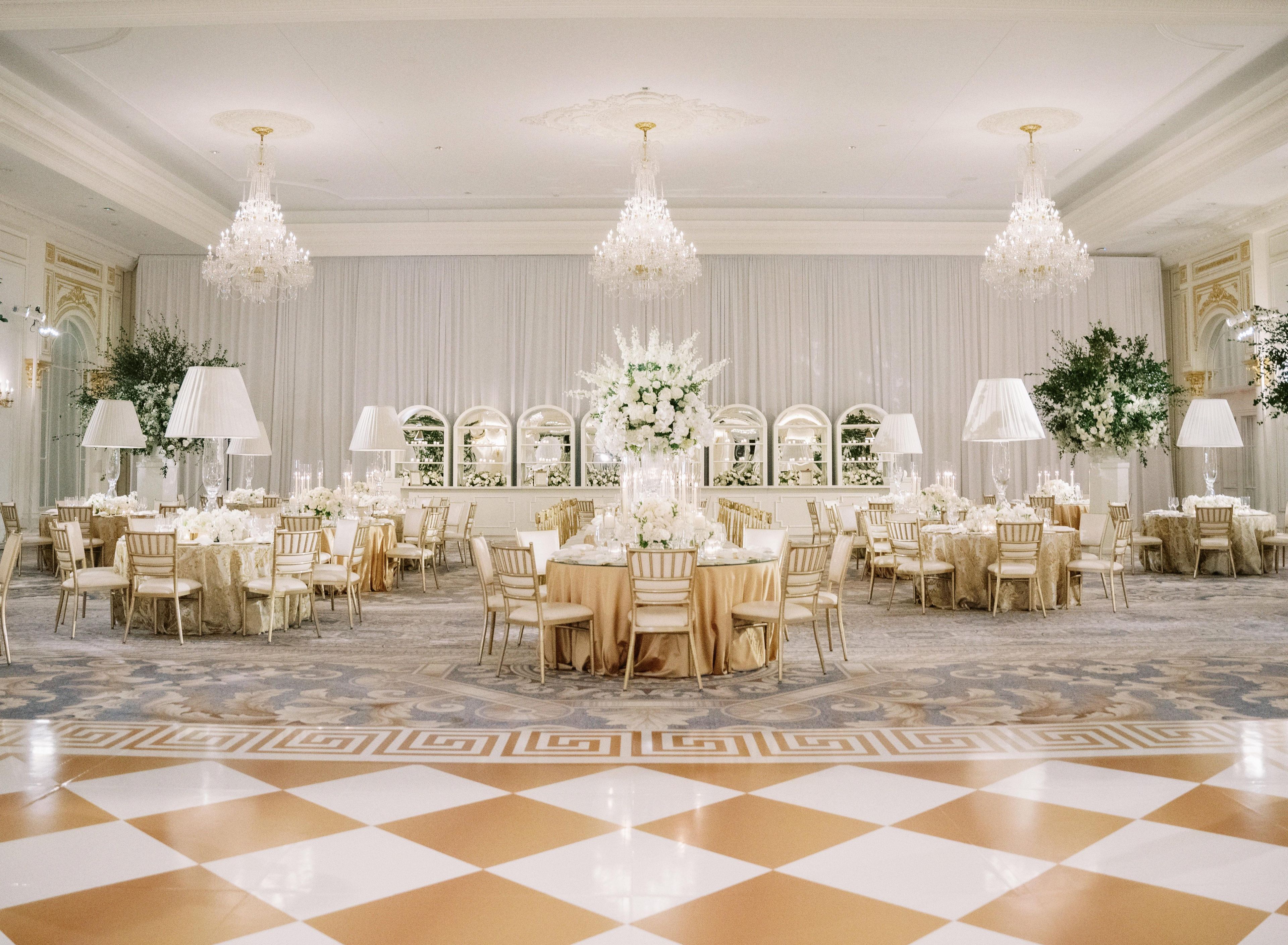 A grand ballroom with multiple round tables, elegant chandeliers, and a checkered floor pattern.