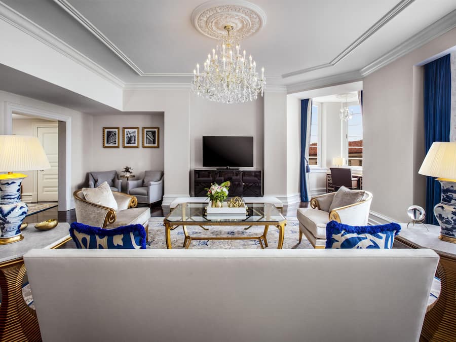 A spacious living room with a chandelier, a flat-screen TV, and a fireplace. There are two sofas, a coffee table, and decorative lamps.