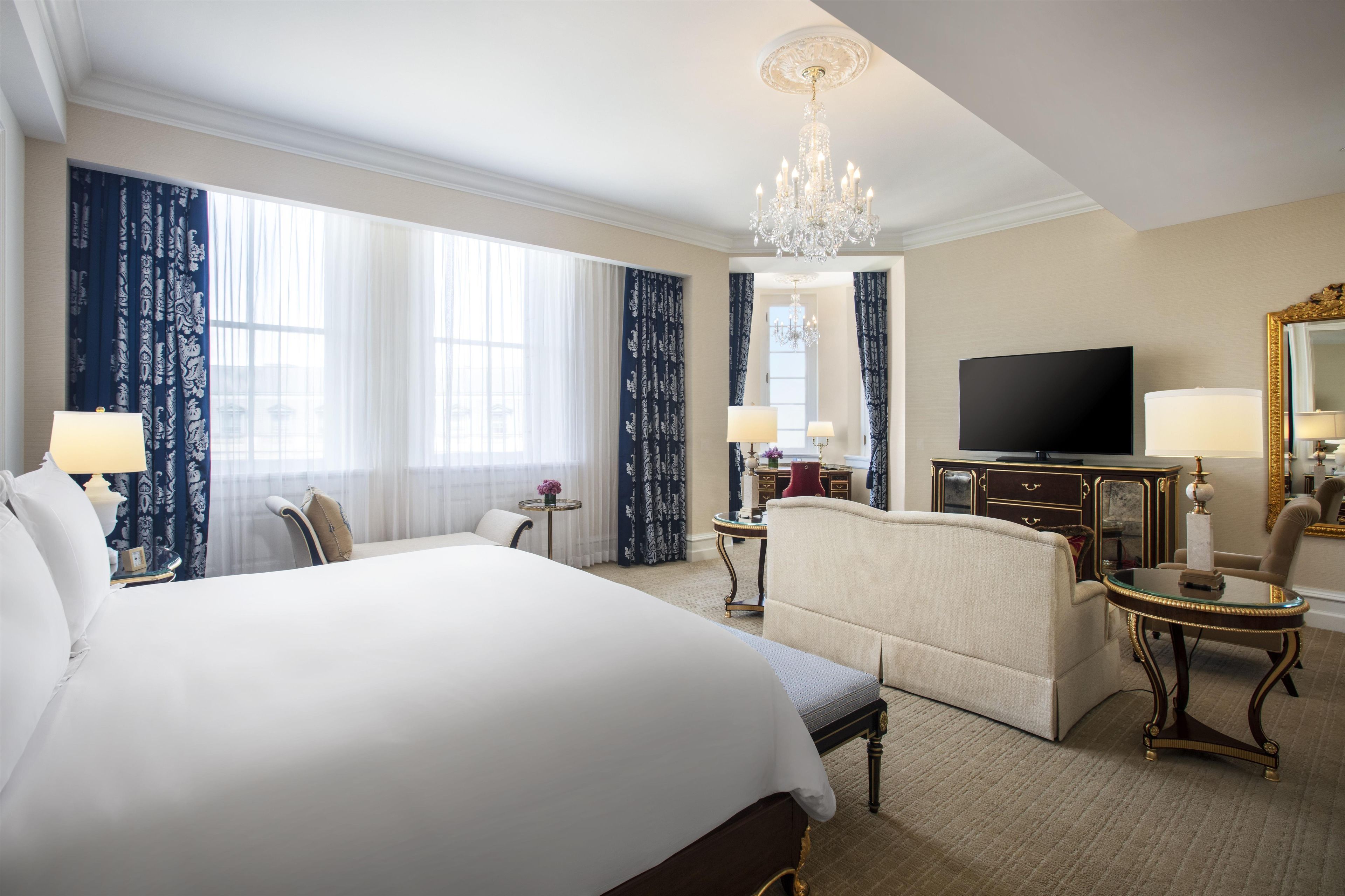 A hotel room with a large bed, a sofa, a small table, a TV, and a chandelier.