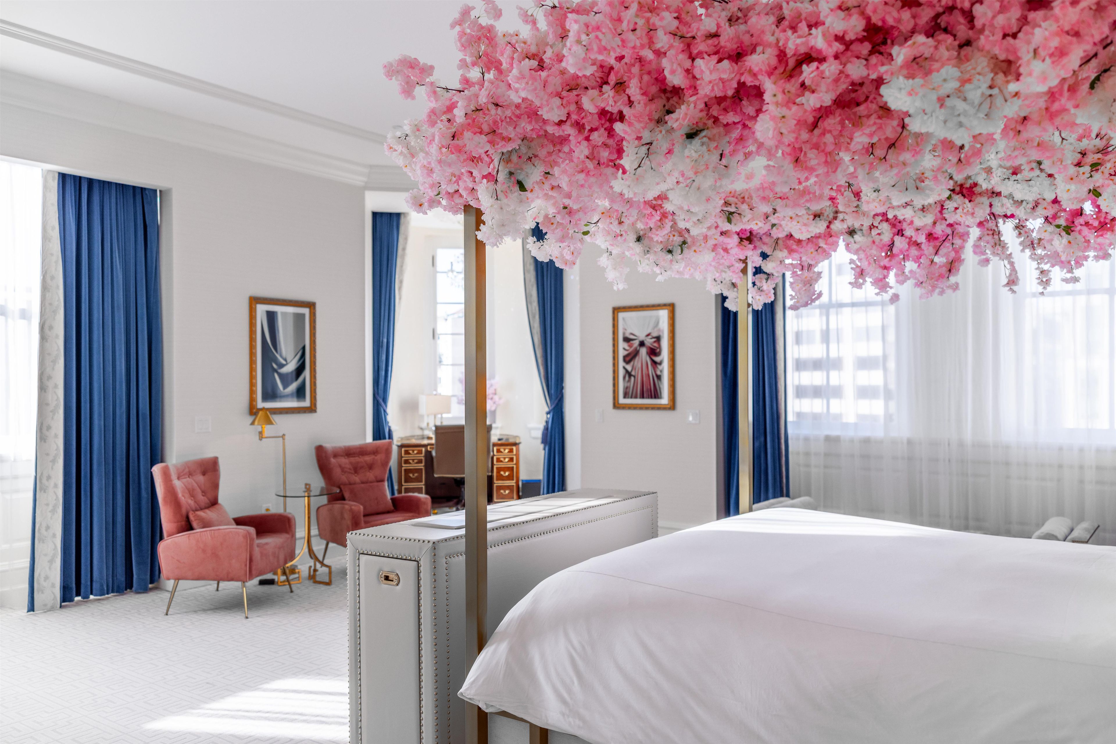 A bedroom with a large bed, a desk, two chairs, and a decorative tree with pink flowers.