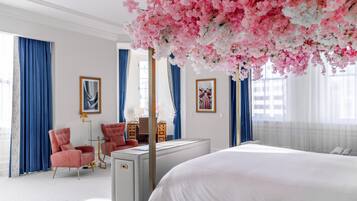 A bedroom with a large bed, a desk, two chairs, and a decorative tree with pink flowers.