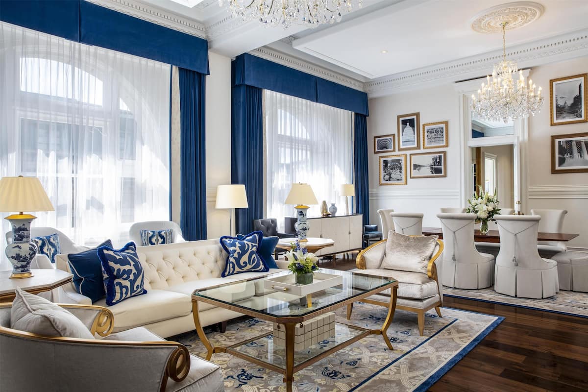 Ein geräumiges Wohnzimmer mit einem weißen Sofa, blauen und weißen Kissen, einem Glastisch und einer Kristallleuchtreiter.