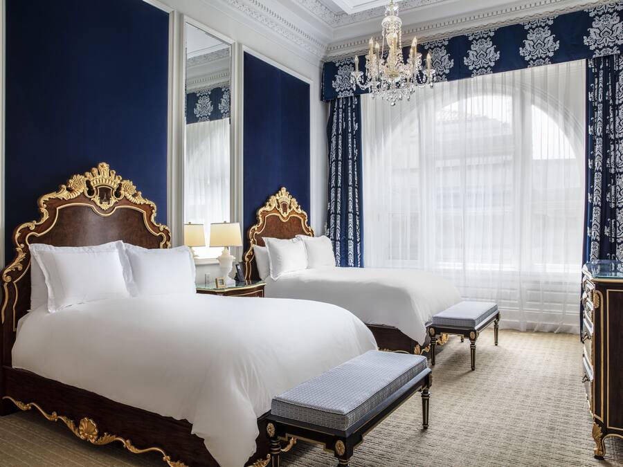 A hotel room with two large beds, a bench, a dresser, and a chandelier.