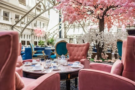A round table set with white teacups and saucers, a teapot, and a tray, surrounded by pink upholstered chairs.