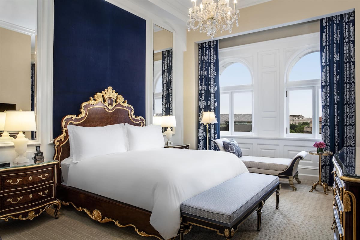 Ein luxuriöses Hotelzimmer mit einem großen Bett, einem aufwendigen Kopfteil und Blick auf die Stadt durch große Fenster.