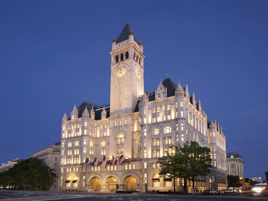 A large, historic building with a prominent clock tower, multiple arches, and flags.