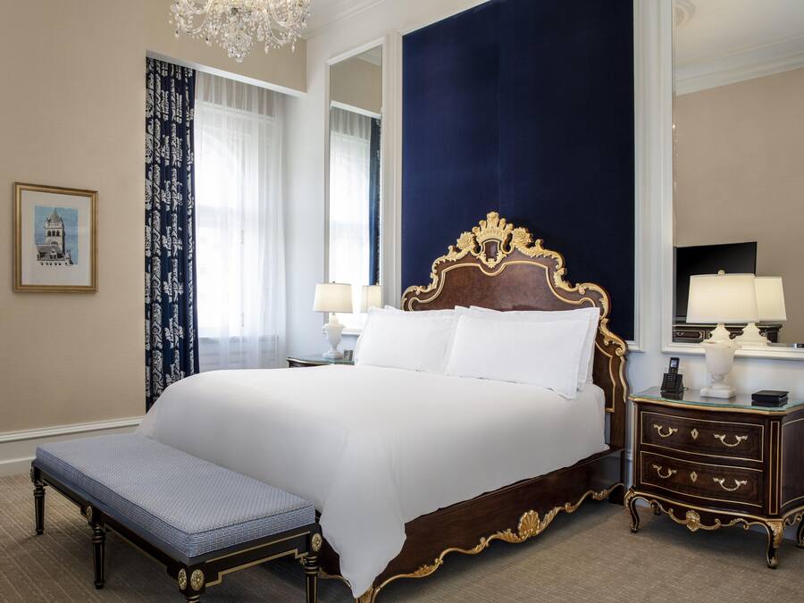 Uma cama grande com cabeceira de madeira, uma mesa de cabeceira com luminárias e um banco.