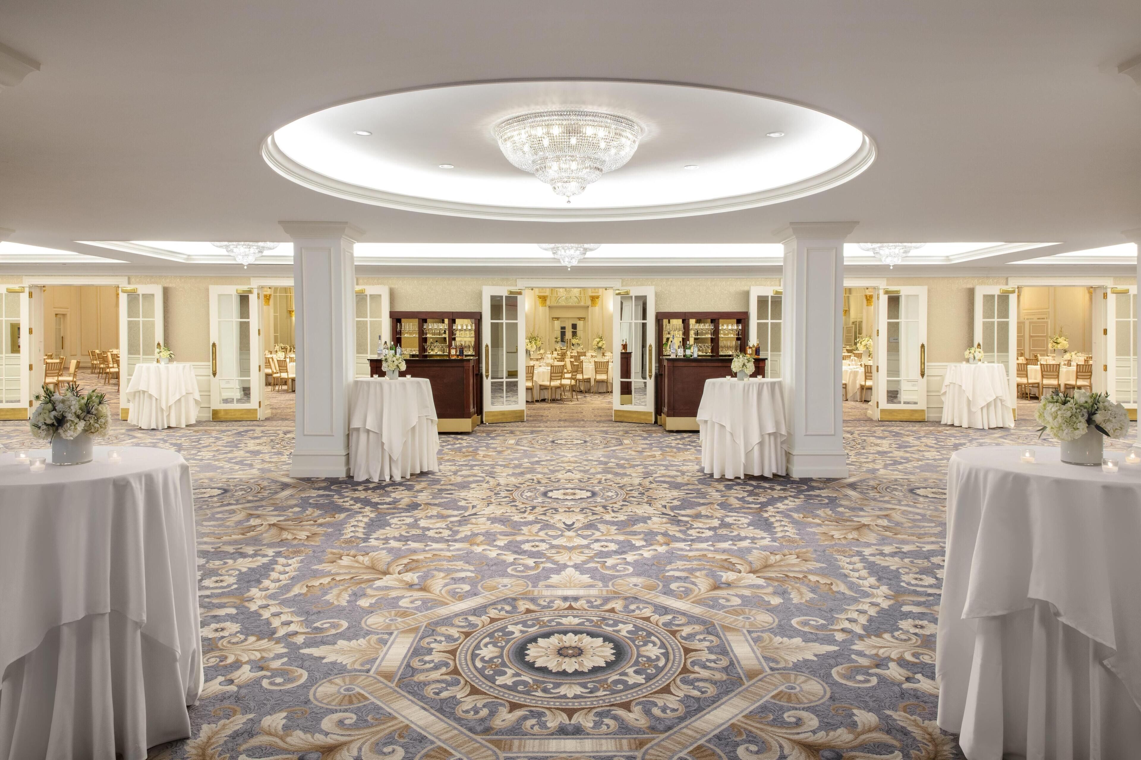 A grand hall with ornate carpet, chandeliers, and multiple tables set for dining.
