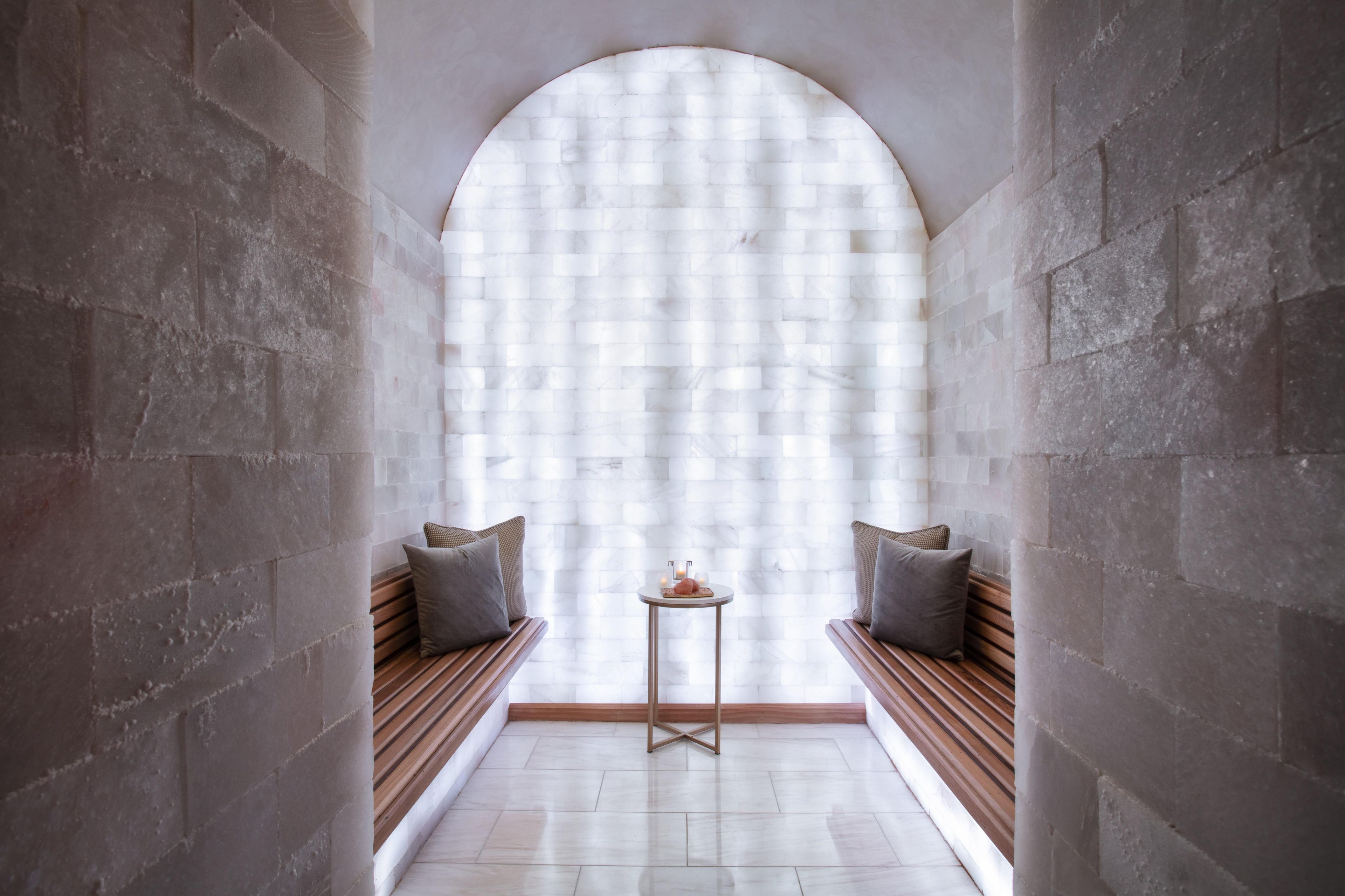 A narrow hallway with a large arched window, two wooden benches with cushions, and a small round table with a candle.
