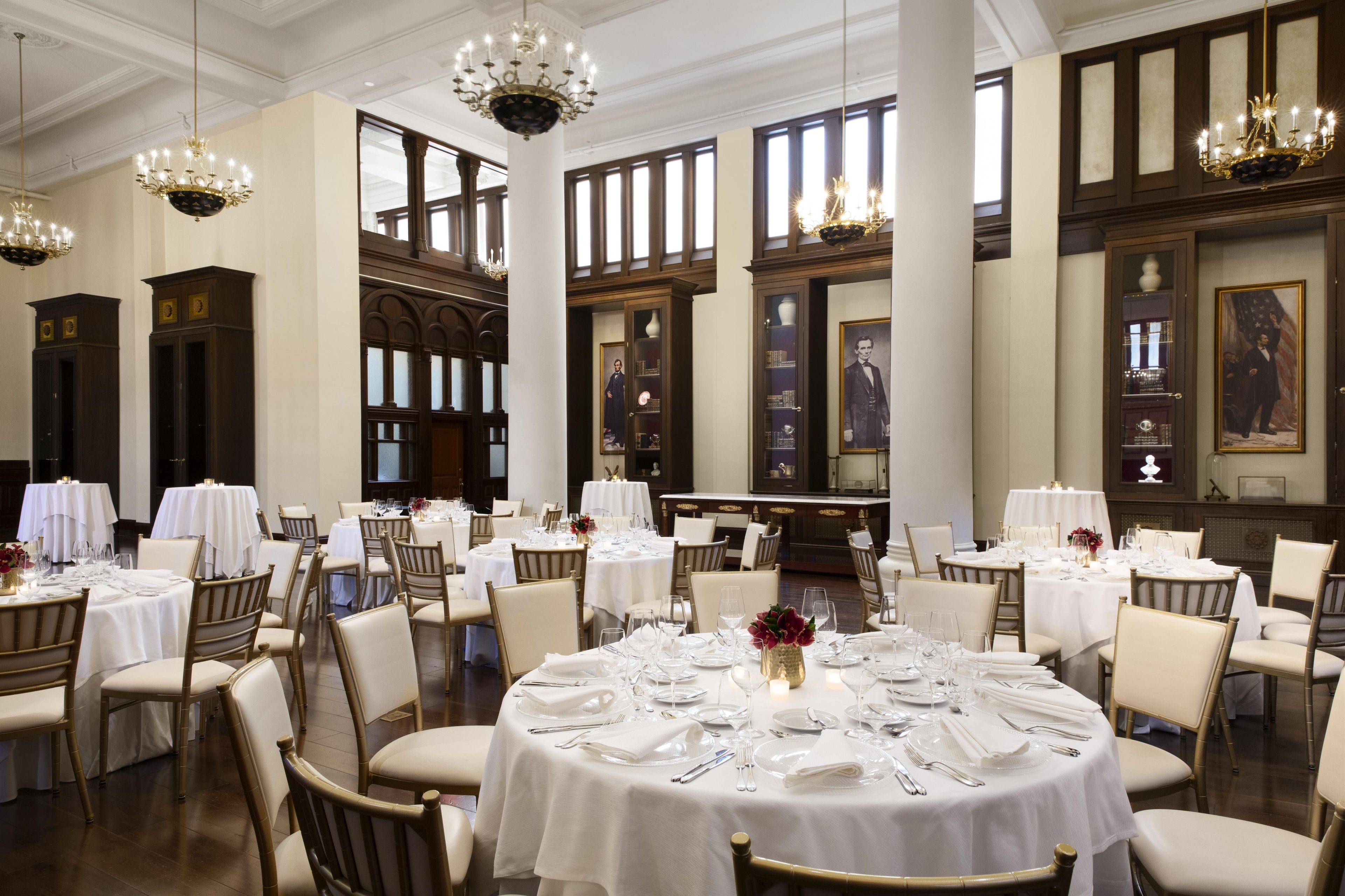 Uma sala de jantar formal com mesas redondas preparadas para o serviço, grandes janelas e painéis de madeira clássicos.