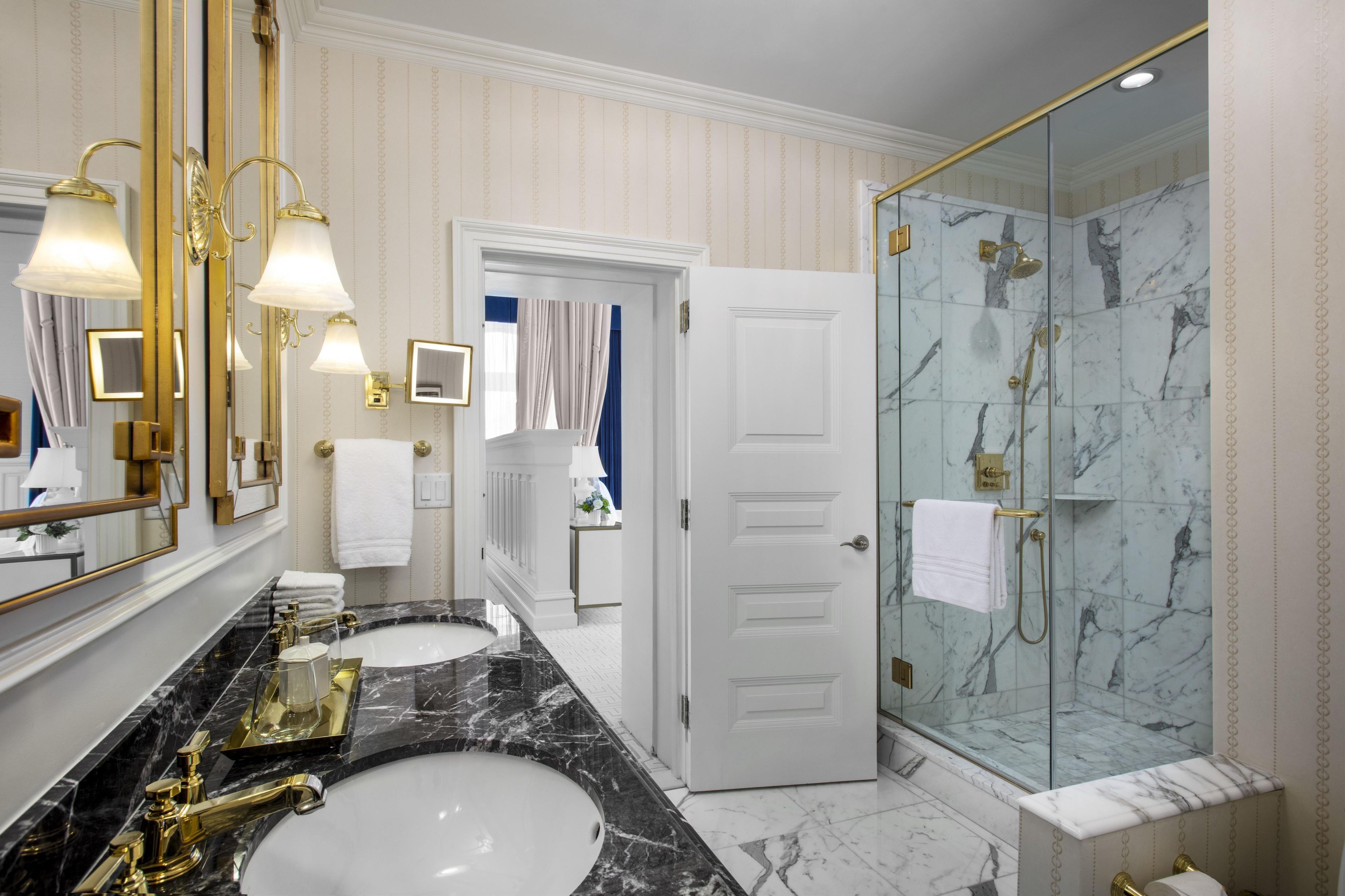 A luxurious bathroom with two sinks, a glass-enclosed shower, and a large mirror with gold fixtures.