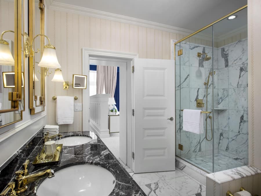 A luxurious bathroom with two sinks, a glass-enclosed shower, and a large mirror with gold fixtures.