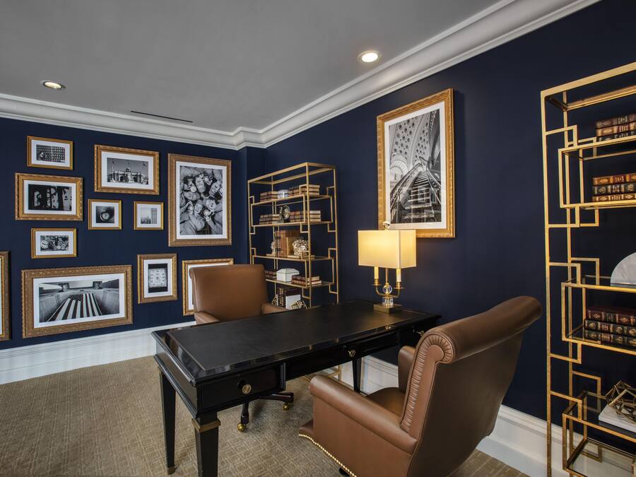 A room with a dark blue wall, a black desk, two brown leather chairs, and a gold-framed bookshelf.
