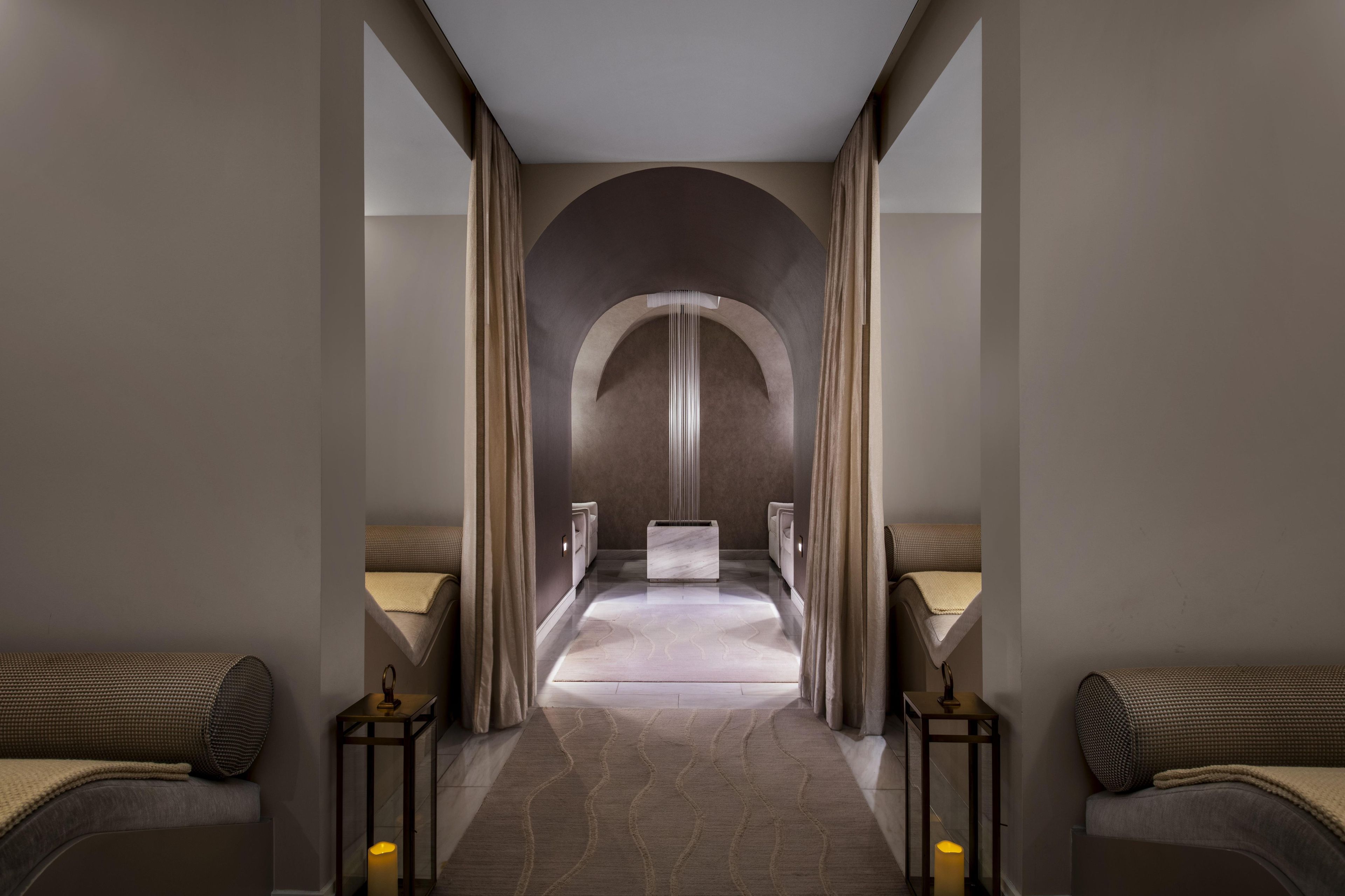 A modern hotel corridor with a central arched doorway, two side beds, and a marble floor.