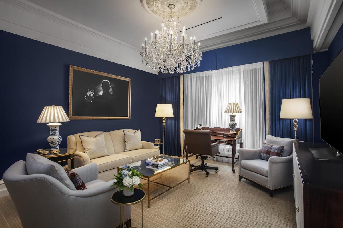 Ein modernes Wohnzimmer mit einem beigen Sofa, einem Glastisch und einem Schreibtisch mit Stuhl. Es gibt eine Deckenleuchte, einen Fernseher und ein großes Fenster mit Vorhängen.