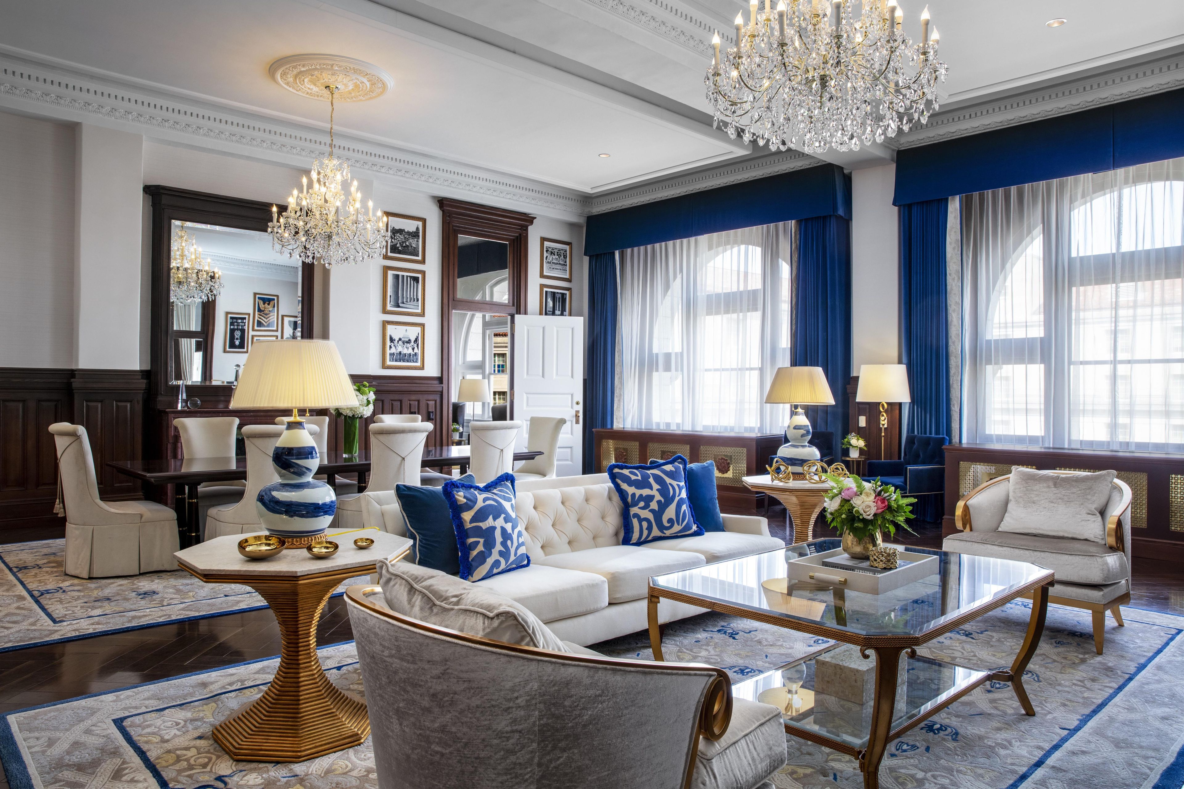 Uma sala de estar luxuosa com um sofá branco, almofadas estampadas em azul e branco, uma mesa de centro de vidro e um lustre.