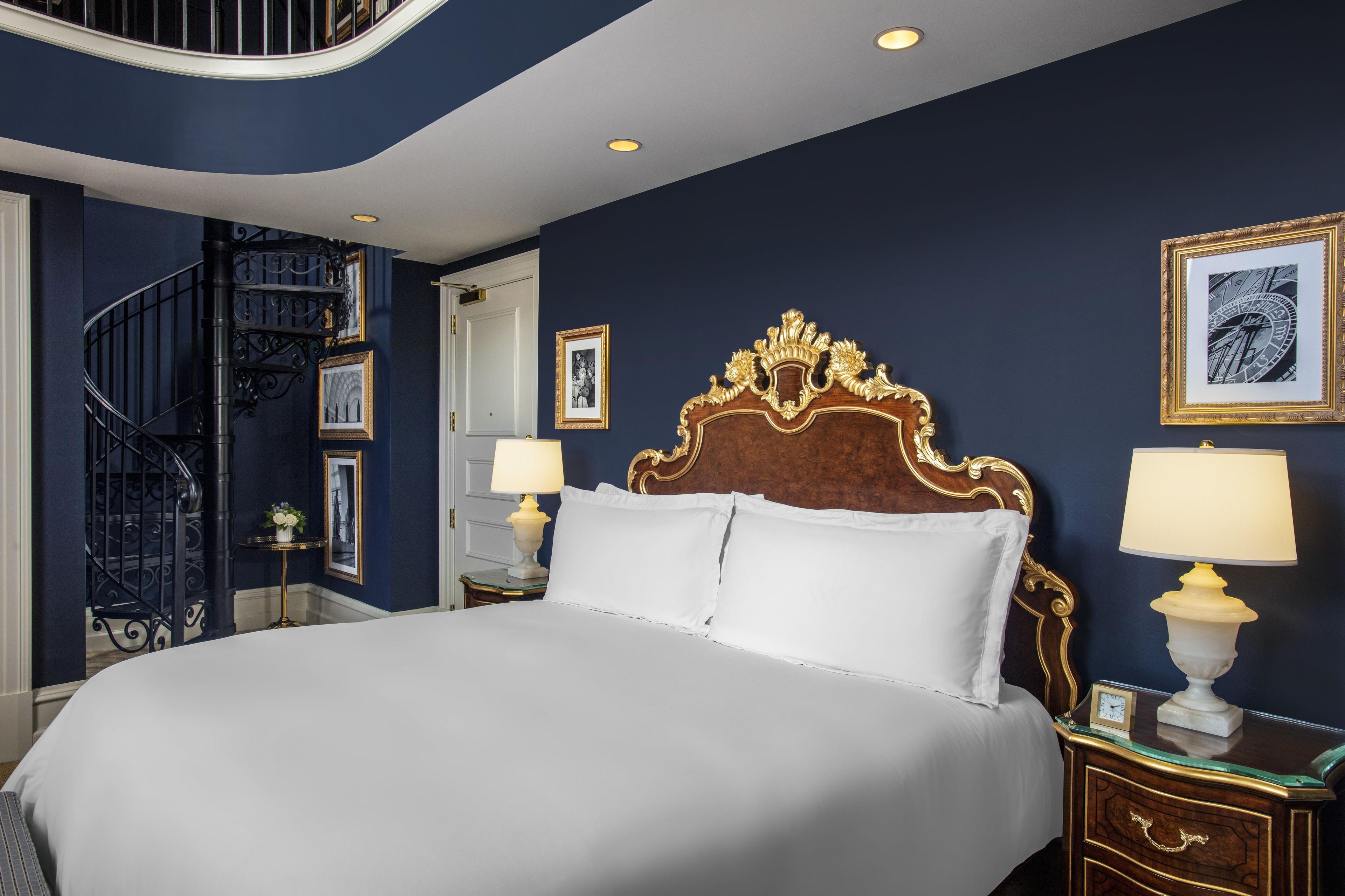 A bedroom with a large bed, a wooden headboard, two bedside tables with lamps, a small clock, and a framed picture on the wall.