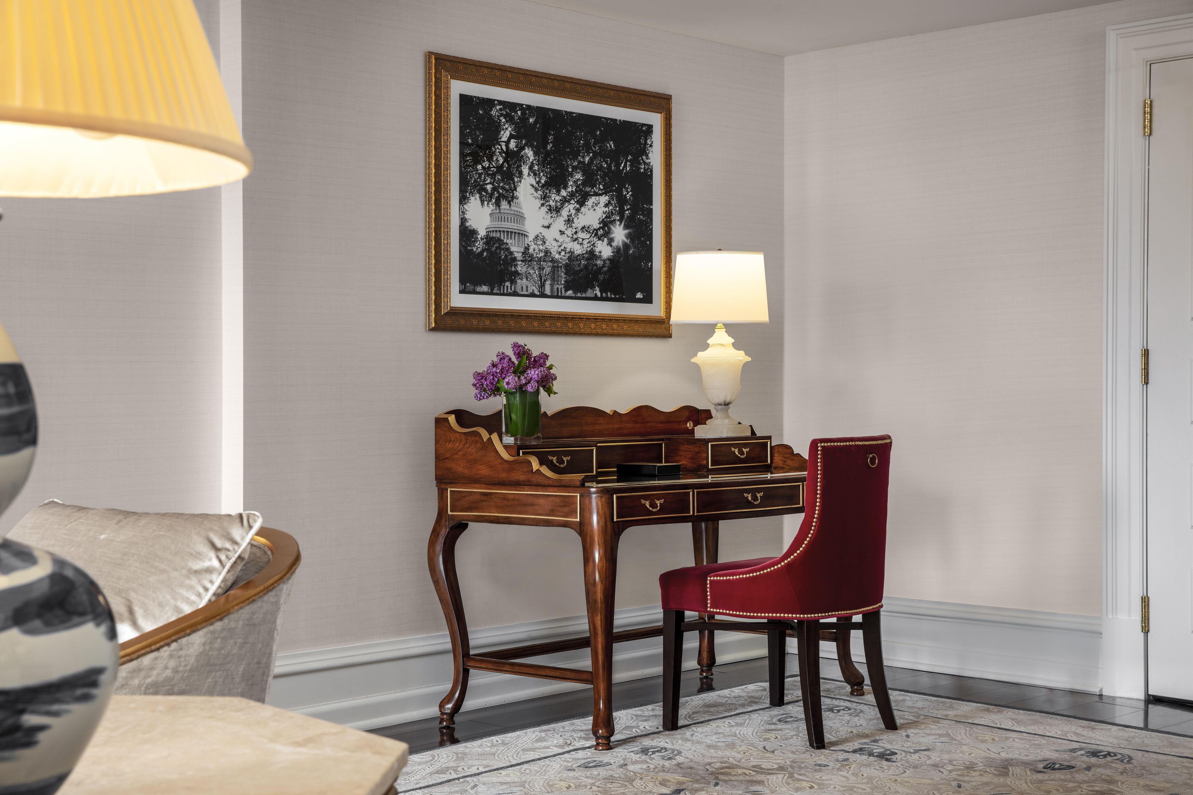 Quarto com escrivaninha de madeira, cadeira vermelha, luminária, quadro pendurado na parede e vaso com flores.