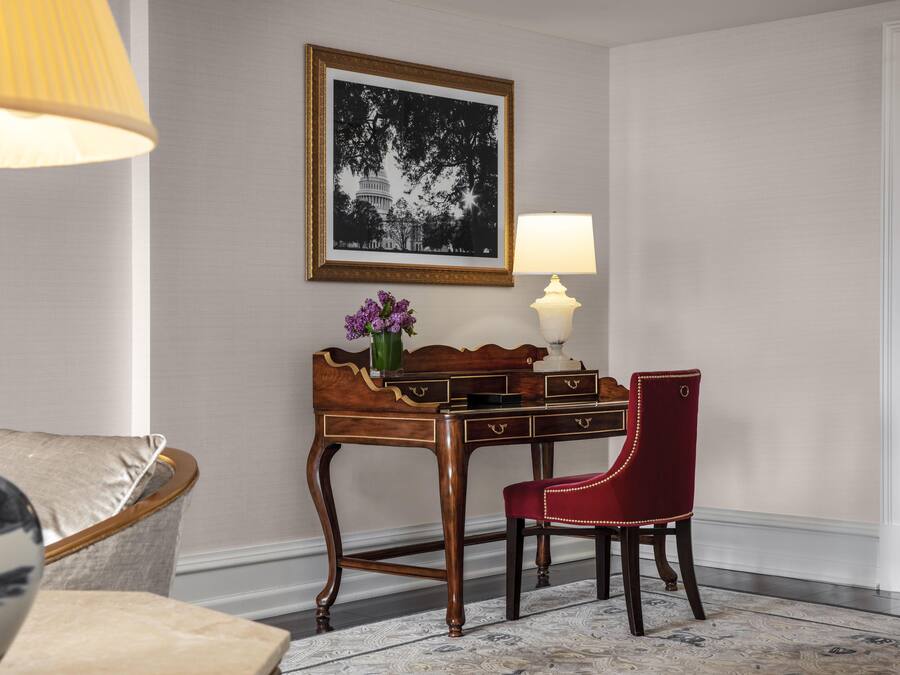 Quarto com escrivaninha de madeira, cadeira vermelha, luminária, quadro pendurado na parede e vaso com flores.