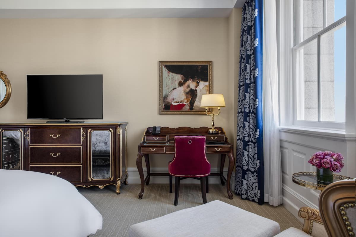 Ein Hotelzimmer mit einem Bett, einem Schreibtisch mit Stuhl, einem Fernseher, einem Gemälde, einer Lampe und einem Fenster mit Vorhängen.