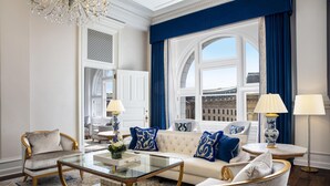 A luxurious living room with a white sofa, blue and gold accents, a glass coffee table, and a view of the city through large windows. - Waldorf Astoria Washington DC (Washington)