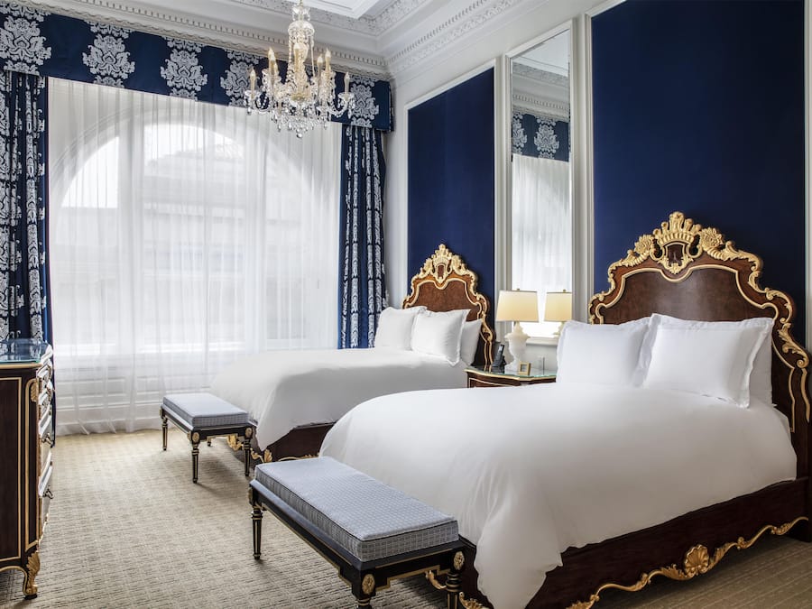 Une chambre d’hôtel avec deux lits, un lustre, un grand miroir et une commode en bois classique.
