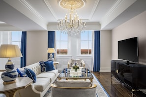 A spacious living room with a chandelier, a sofa with blue cushions, a coffee table, a television, and a large window with curtains.