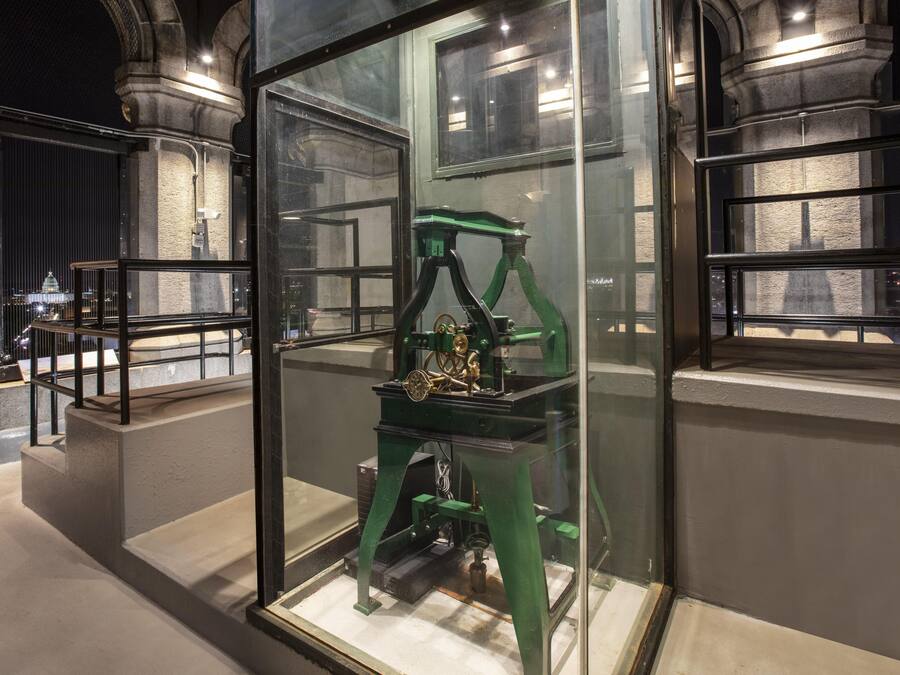 Una exhibición de museo con un dispositivo mecánico dentro de una vitrina, rodeada de arquitectura de piedra.